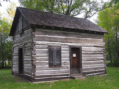A full shot of the Bergquist Cabin in north Moorhead. The oldest building in the community on its original site, the cabin is seen from the southwest corner, showing two doors and two windows set in the gray log walls.